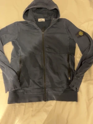 Stone Island hoodie - Säljer en blå hoodie från Stone Island med dragkedja framtill och två fickor med dragkedja. Klassisk patch med logga på vänster ärm. Hoodien har huva och ribbade muddar. Perfekt för en avslappnad streetwear-look.