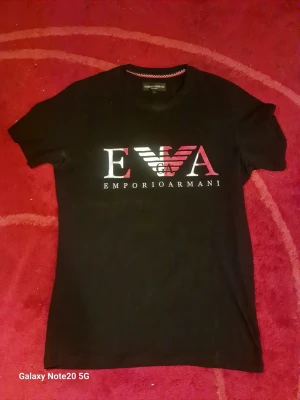 Svart Emporio Armani t-shirt - Snygg svart t-shirt från Emporio Armani med stort logotryck i vitt och rosa på bröstet. Klassisk rund halsringning och korta ärmar. Perfekt för dig som gillar stilrena och trendiga plagg med tydlig branding.