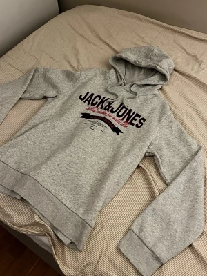 Grå hoodie från Jack & Jones - Snygg grå hoodie från Jack & Jones med tryck i svart och rött på bröstet. Klassisk huva med dragsko och ribbade muddar vid ärmar och nederkant. Perfekt för en avslappnad och trendig stil. Tillverkad i mjukt material för extra komfort.
