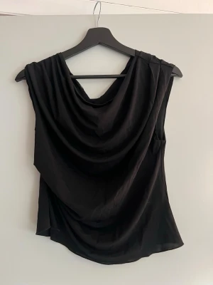 Off-shoulder topp - Snygg svart topp med draperad front och ärmlös design. Toppen har en lös passform och ett mjukt, följsamt material som ger ett elegant fall. Perfekt att styla med jeans eller kjol för en trendig look.