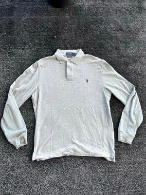 Ralph Lauren pike - Ljusgrå långärmad pikétröja från Polo Ralph Lauren med klassisk krage och knäppning framtill. Tröjan har den ikoniska lilla logotypen broderad på bröstet och är tillverkad i mjukt bomullsmaterial. Perfekt för en clean och stilren look. Storlek L, skick 9/10. Pris går att diskutera✅