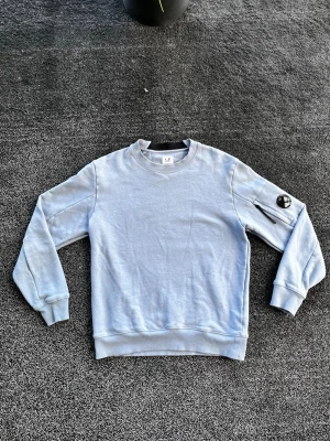 C.P. Company sweatshirt - Ljusblå sweatshirt från C.P. Company med rund halsringning och ribbade muddar. Tröjan har en ikonisk detalj med svart lins på ärmen och en liten ficka med dragkedja. Perfekt för en avslappnad och stilren look. Storlek M, äkta, skick 9/10. Pris går att diskutera✅