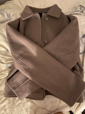 Brun ullkappa med stora knappar - Stilren brun kappa i ullmix med klassisk krage och stora knappar framtill. Kappan har raka ärmar och diskreta fickor på sidorna. Perfekt för kyligare dagar och ger en clean look till din outfit. kommer ej till användning och ligger bara o tar plats, helt ny använt en gång 🤍