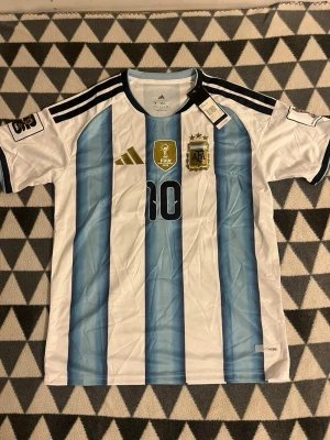 Argentina matchtröja nr 10 Adidas - Säljer en officiell Argentina fotbollströja från Adidas med nummer 10, klassiska blå och vita ränder, guldiga detaljer och FIFA World Champions 2022-märke. Tröjan har korta ärmar och broderade loggor. Perfekt för fotbollsfans!
