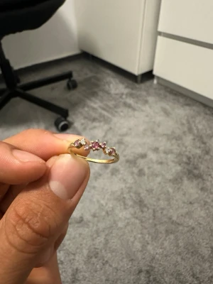 Guldring med rosa och vita stenar - Snygg guldfärgad ring med fem glittrande stenar i rosa och vitt. Stenarna är runda och sitter i rad på ovansidan av ringen, vilket ger en elegant och trendig look. Perfekt accessoar för att lyfta din outfit med lite extra färg och bling.
