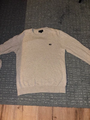Beige rundhalsad tröja - Snygg beige tröja med rund halsringning och diskret logga på bröstet. Tröjan har långa ärmar och ribbade muddar vid ärmslut och nederkant. Perfekt för en stilren och enkel look.