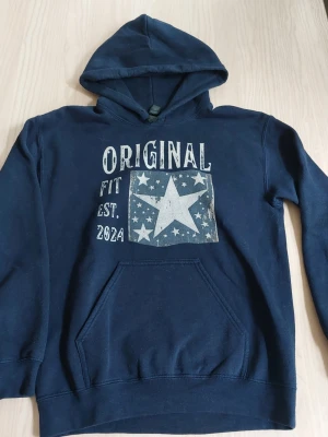 Mörkblå hoodie med stjärntryck - Snygg mörkblå hoodie med huva och magficka. Tryck på bröstet med texten 'ORIGINAL FIT EST. 2024' och en stor vit stjärna omgiven av mindre stjärnor. Perfekt för en avslappnad och cool stil.