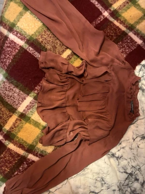 Rostbrun höstig blus med volanger  - Säljer en snygg rostbrun blus med långa ärmar och flera volangdetaljer både fram och bak. Blusen har en rund halsringning och är gjord i ett tunt, lätt material som ger ett flowy intryck. Perfekt för dig som gillar romantiska detaljer och en trendig look. Höstig och lite genomskinlig 