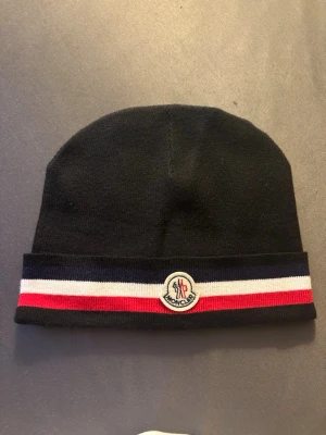Svart Moncler mössa med ränder - Snygg svart mössa från Moncler med klassiska ränder i blått, vitt och rött längs kanten. Mössan har Moncler-logga framtill och är stickad i mjukt material som håller dig varm och stilren under kalla dagar.