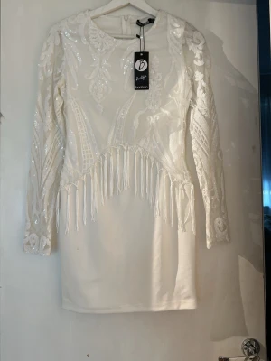 Vit paljettklänning med fransar Boohoo - Vit kort klänning från Boohoo med långa ärmar, dekorerad med paljetter och broderade mönster. Klänningen har fransdetaljer framtill och en rund halsringning. Perfekt för dig som vill sticka ut med en unik look.