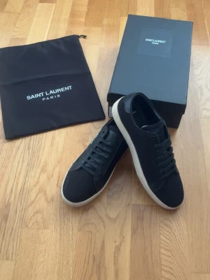 Saint Laurent skor - Snygga svarta sneakers från Saint Laurent med vit sula och svart ovandel i canvas. Klassisk låg modell med svarta skosnören och diskret Saint Laurent-logga på sidan. Perfekta för en stilren och trendig look. Pris kan diskuteras vid snabb affär!