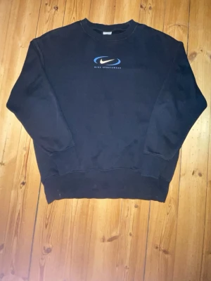 Svart sweatshirt från Nike - Klassisk svart sweatshirt från Nike med blått och vitt tryck på bröstet där det står 'Nike Sportswear'. Tröjan har rund halsringning, ribbade muddar vid ärmslut och nederkant. Perfekt för en avslappnad och sportig stil.