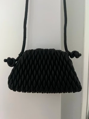 Svart axelremsväska med flätad look - Snygg svart handväska med unik flätad struktur och axelrem. Väskan har en mjuk, rundad form och knutdetaljer på remmen. Perfekt för dig som vill ha en trendig accessoar med cool textur. Materialet ser ut att vara konstläder eller liknande.