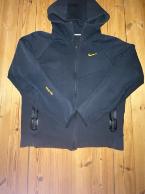 Nike NOCTA hoodie med dragkedja - Svart hoodie från Nike NOCTA med gul logga på bröstet och NOCTA-tryck på ärmen. Tröjan har dragkedja framtill, två fickor med dragkedja och en stor huva. Materialet ser ut att vara mjukt och bekvämt, perfekt för en chill och sportig look.