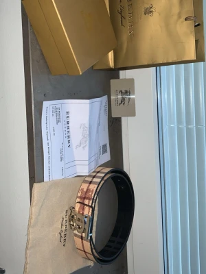 Burberry rutigt bälte med guldspänne - Snyggt bälte från Burberry med klassiskt rutigt mönster i beige, brunt och svart. Bältet har ett elegant silver spänne med Burberry-logga och är tillverkat i skinn. Perfekt accessoar för att lyfta din outfit. PSA! man kan ta bort burberry silver spännet och lägga den på andra sidan så att man får en svart burberry bälte också.