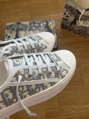 Dior gråvita låga sneakers - Snygga låga sneakers från Dior i grått och vitt med det klassiska Dior-mönstret på canvas och vita detaljer. Skorna har vita snören, gummisula och logga på plösen. Perfekta för dig som vill ha en exklusiv och trendig look.