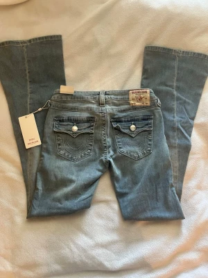 Ljusblå True Religion bootcut jeans - Säljer ett par ljusblå bootcut jeans från True Religion med ikoniska fickor bak och vita kontrastsömmar. Jeansen har dekorativa knappar på ficklocken och klassisk patch bak i midjan. Perfekta för dig som gillar en avslappnad men snygg stil. Modellen heter JOEY low rise flare 🌟🌟 storleken är W27, Innerbenslängd: 84cm 🔥 de är helt nya och aldrig använda, nypris är 1400kr