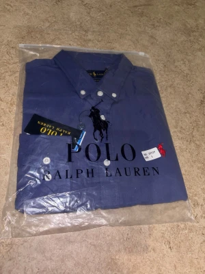 Blå skjorta från Polo Ralph Lauren - Klassisk blå skjorta från Polo Ralph Lauren med knappslå och vit knappdetalj. Skjortan har en broderad röd logga på bröstet och är tillverkad i bomull. Perfekt för en stilren och tidlös look.