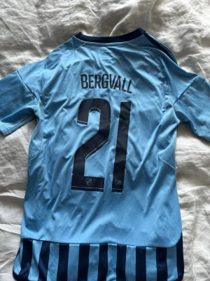 Djurgården matchtröja Bergvall #21 - Säljer en ljusblå och marinblå randig Djurgården matchtröja från Adidas med namn och nummer Bergvall 21 på ryggen. Tröjan har klubbmärke på bröstet, korta ärmar och klassiska Adidas-ränder på axlarna. Perfekt för dig som älskar fotboll och Djurgården. Storlek är 13/14 men passar även xs-xs