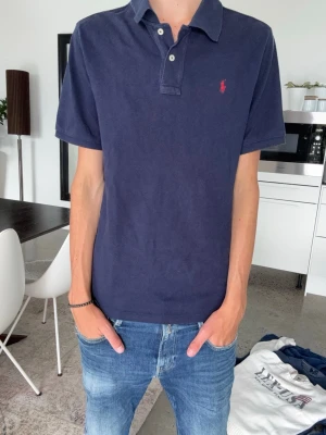 Mörkblå piké från polo Ralph Lauren - Skön marinblå piké t-shirt med klassisk krage och två knappar framtill. T-shirten har korta ärmar och en liten röd broderad logga på bröstet. Tillverkad i mjuk bomull som känns skön mot huden. Perfekt till jeans eller shorts för en clean look. Barn XL 18-20 passar som en större S