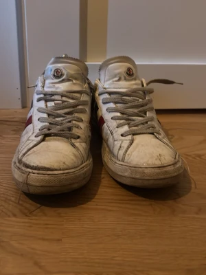Vita Moncler sneakers med röd/blå detalj - Säljer ett par vita Moncler sneakers med grå skosnören och klassiska röd och blå ränder på sidan. Skorna har låg profil och rund tå, samt Moncler-logga på plösen. Tillverkade i läder med gummisula. Perfekta för dig som gillar streetstyle och exklusiva märken.