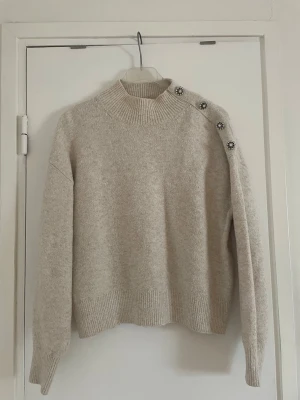 Beige stickad tröja - Säljer en beige stickad tröja med ribbad hög hals och långa ärmar. Tröjan har fyra dekorativa pärlknappar längs ena axeln som ger en unik touch. 