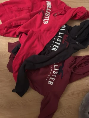 Tre Hollister hoodies i olika färger - Säljer tre snygga hoodies från Hollister i färgerna röd, vinröd och svart. Alla har stor logga och text på bröstet samt klassisk huva och känguruficka. Perfekta för en chill och avslappnad stil. Materialet känns mjukt och skönt.