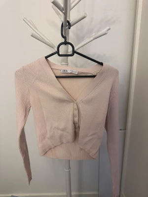 Ljusbeige ribbad cardigan från ZARA - Söt ljusrosa ribbad cardigan från ZARA med v-ringning och knappar framtill. Croppad modell med långa ärmar, perfekt att styla till både jeans och kjol. Mjuk och stretchig känsla i materialet.