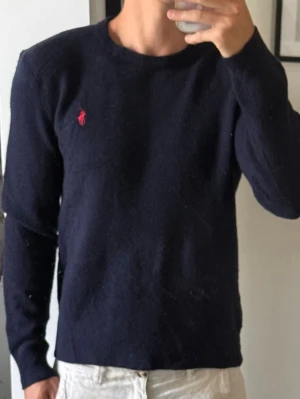 Mörkblå stickad tröja Polo Ralph Lauren - Klassisk mörkblå stickad tröja från Polo Ralph Lauren med rund halsringning och röd broderad logga på bröstet. Tröjan har långa ärmar och ribbade muddar vid ärmslut och nederkant. Perfekt till jeans eller chinos. M men är liten i storleken så sitter som S. 100% merinoull 