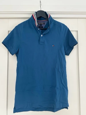 Blå pikétröja med randig krage - Snygg blå pikétröja med korta ärmar och klassisk krage med röd, vit och blå rand. Liten logga på bröstet och knappar vid halsen. Perfekt till jeans eller shorts för en clean look.