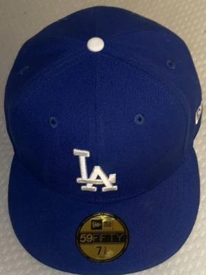 Blå LA 59FIFTY keps från New Era - Snygg blå 59FIFTY keps från New Era med LA-logga broderad framtill i vitt. Klassisk flat brim och ventilationhål runt om. Kepsen har en vit knapp på toppen och sticker ut med sin sportiga look. Perfekt för dig som gillar streetwear och vill ha en ikonisk accessoar.   58.7cm OMKRETS