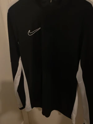 Svart och vit Nike träningströja - Snygg svart långärmad träningströja från Nike med vita detaljer längs ärmar och sidor. Tröjan har en hög krage och dragkedja upptill. Perfekt för träning eller uppvärmning. Materialet är troligtvis polyester för bra andningsförmåga.