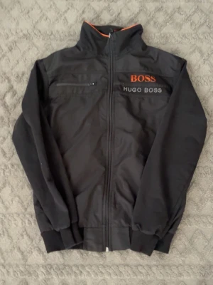 Svart vindjacka från Hugo Boss - Snygg svart vindjacka från Hugo Boss med dragkedja framtill och en bröstficka med zip. Jackan har hög krage med orange detalj och broderad logga på bröstet. Perfekt för dig som vill ha en stilren och sportig look.