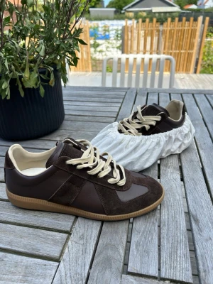 Bruna gats, margiela - Snygga bruna sneakers från Maison Margiela med beige snörning och detaljer i mocka och läder. Skorna har en klassisk låg siluett och beige yttersula. Perfekta för dig som gillar stilrena och tidlösa sneakers. Säljer dem eftersom att dem är 1 storlek för liten. Detta är mest än intressekoll! Har ett par vita också i storlek 42 om någon är intresserad. Har ingen stress med att sälja isåfall ger jag dem bara till min bror!