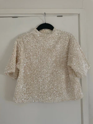 Paljettprydd topp - Säljer en glittrig beige/vit topp täckt med paljetter. Toppen har korta ärmar, rak passform och hög hals. 