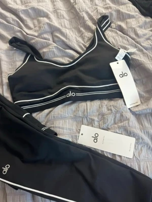 Svart träningsset från Alo med vita detaljer - Snyggt svart träningsset från Alo med sport-bh och tights. Setet har vita kontrastlinjer och logga på både bh och byxa. Materialet är stretchigt och följsamt, perfekt för gym eller yoga. Sport-bhn har smala axelband och tightsen har hög midja.