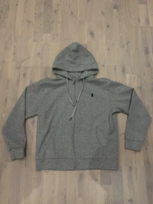 Grå zip hoodie från Polo Ralph Lauren - Säljer en grå hoodie med dragkedja från Polo Ralph Lauren. Klassisk design med huva och dragsnören, två fickor framtill och den ikoniska svarta loggan broderad på bröstet. Perfekt för en chill och stilren look.