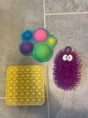 Fidgetleksaker: pop it och stressboll - Tre olika fidgetleksaker säljes: en fyrkantig gul pop it, en färgglad pop it i form av en tass och en lila taggig stressboll med ögon. Perfekt för att sysselsätta händerna eller bara ha kul med kompisar.