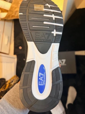 Svarta sneakers från Emporio Armani - Stilrena svarta sneakers från Emporio Armani med vita och blå detaljer. Skorna har tydlig EA7-logga på sidan och sulan, samt snörning framtill. Ovandelen är i ett ventilerande syntetmaterial och sulan är robust med bra grepp.