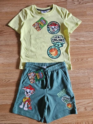Paw Patrol T-shirt och shorts set - Snygg gul t-shirt med Paw Patrol-tryck och Dino Island-motiv. T-shirten har rund hals, korta ärmar och färgglada tryck framtill med Marshall och dinosaurietema. Perfekt för dig som gillar tecknade serier och vill ha en lekfull stil. Med tryck från nya filmen som snart har premiär!  Tröjan är 100% bomull och shortsen är 80% bomull och 20% polyester.