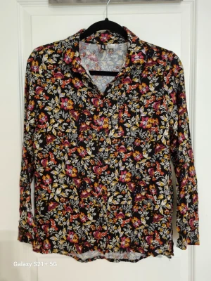 Blommig svart blus med krage - Snygg svart blus med färgglatt blommönster i rött, gult, vitt och lila. Blusen har krage, knäppning framtill och långa ärmar. Materialet känns mjukt och lätt, perfekt för en trendig look. Passar till både jeans och kjol.