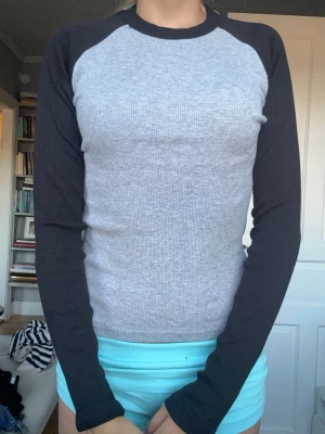 Grå långärmad baseballtröja - Säljer en grå långärmad baseballtröja med svarta ärmar och svart krage. Tröjan har en ribbad struktur och en klassisk rak passform. Perfekt för en avslappnad och sportig stil.