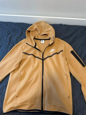 Nike tech tracksuit Camel (ljus kamelbrun) - Snygg beige hoodie från Nike med hel dragkedja och svart detalj längs bröstet och ärmarna. Hoodien har huva, två sidofickor och en extra ficka med dragkedja på ena ärmen. Tillverkad i mjukt material som känns skönt mot huden. Perfekt för en avslappnad streetwear-look.