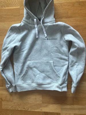 Grå hoodie från Polo Ralph Lauren - Snygg grå hoodie från Polo Ralph Lauren med vit dragsko och klassisk logga broderad på bröstet. Hoodien har känguruficka framtill, ribbade muddar och är tillverkad i mjukt sweatshirtmaterial. Perfekt för en chill och stilren look.