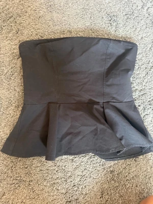 Svart axelbandslös peplum topp - Säljer en svart axelbandslös topp med peplumdetalj nedtill. Toppen har en figurnära passform upptill och en utsvängd nederdel som ger en snygg siluett. (Säljer likadan i ljusblå)