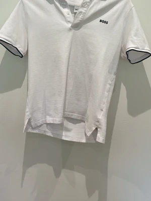 Vit pikétröja från BOSS XS - Vit pikétröja från BOSS med svart logga på bröstet och svarta detaljer vid ärmsluten. Klassisk krage med knappar och slitsar i sidorna. Perfekt för en clean och stilren look.
