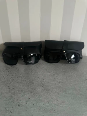Svarta solglasögon med fodral - Två par svarta solglasögon med rektangulära bågar och mörka glas. Båda kommer med matchande svarta fodral med knappstängning. Snygg och stilren design som passar till många outfits.