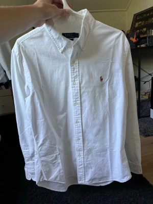 Vit skjorta från Polo Ralph Lauren - Klassisk vit skjorta från Polo Ralph Lauren med button-down krage och broderad logga på bröstet. Skjortan har lång ärm, normal passform och är tillverkad i mjuk bomull. Perfekt till jeans eller chinos för en clean look.