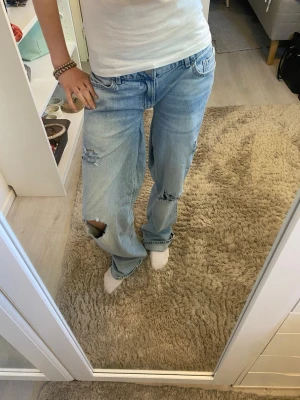 Ljusblå slitna jeans från Lindex - Säljer ett par ljusblå jeans från Lindex med avslappnad passform och flera slitningar och hål på benen. Jeansen har klassisk femficksmodell och raka ben. Perfekta för en trendig och avslappnad stil. Omsydda för hand som du kan se på tredje bilden