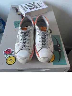 Golden Goose Vit och Orange - Säljer ett par vita sneakers med grå stjärndetalj på sidan och grå snören. Skorna har en orange hälflik och låga sulor. Materialet ser ut att vara läder eller konstläder och designen är klassisk med en modern twist. Storlek 